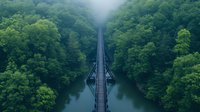Mystical aerial view of symmetrical bridge over calm lake surrounded by lush green forest in foggy morning atmosphere, creating mirror reflection on water surface.