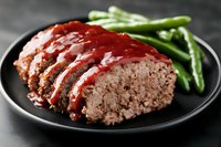 Sliced homemade meatloaf with tangy red glaze sauce served on black plate with steamed green beans. Close up view of comfort food dinner with rich texture.
