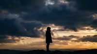 Silhouette of person standing against dramatic cloudy sky at sunset with golden light breaking through storm clouds creating moody atmospheric scene.