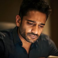 Close-up portrait of South Asian man with concerned expression looking down, illuminated by warm ambient light against dark background.