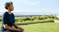 Middle-aged woman with gray hair and dark skin sitting outdoors in a green park, looking toward the horizon with a contemplative expression, wearing a dark blue shirt on a sunny day.