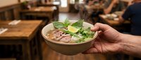 Asian noodle soup with fresh herbs and meat in bowl held by hand in restaurant setting.
