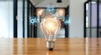 Glowing light bulb with digital innovation icons on wooden desk in modern office space representing creative business ideas and technological solutions.