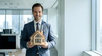 Professional Caucasian businessman in suit holding wooden house model in modern office, real estate investment concept for marketing materials.