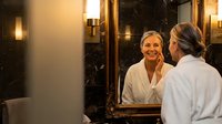 Young Caucasian woman in white bathrobe smiling at her reflection in elegant bathroom mirror with warm lighting for spa and wellness content.