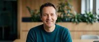 Middle-aged Caucasian man in teal sweater smiling warmly in modern kitchen with wooden cabinets and green plants for lifestyle and home content.
