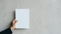 Female hand holding blank white paper sheet against concrete wall background for mockup and branding projects.