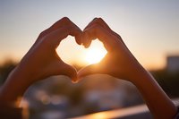 Hands forming heart shape silhouette against golden sunset sky creating romantic love symbol with warm sunlight shining through fingers outdoors.