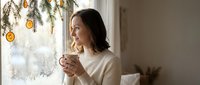 Young woman enjoying hot beverage by window with festive decorations during winter holidays at home for seasonal lifestyle content.