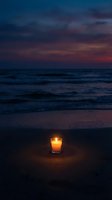 Glowing candle on beach sand at twilight with dramatic purple and orange sunset sky over ocean waves creating peaceful meditation atmosphere.