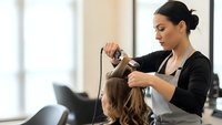 Professional female hairstylist with dark hair in bun styling blonde client's hair with curling iron in modern salon interior.