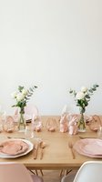 Elegant dining table setting with pink plates, rose gold cutlery, white roses and eucalyptus in glass vases on wooden table for special occasions.