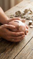 Hands protecting pink piggy bank on wooden table with scattered coins for savings and financial security planning.