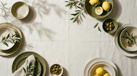Overhead flat lay of Mediterranean cuisine ingredients with olive branches, lemons, and olives arranged on white linen fabric for food styling and culinary design projects.