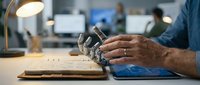 Male engineer or technician with medium skin tone examining circuit board components at workstation with laptop and desk lamp in modern office environment for electronics repair, manufacturing, or technical support purposes.