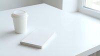 Blank notebook and disposable coffee cup on white desk near window, minimalist workspace setup for business, creative projects, and office branding mockups.
