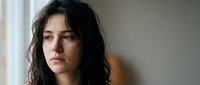 Young brunette woman with wavy hair looking pensively out window in soft natural light. Contemplative mood for mental health awareness campaigns.