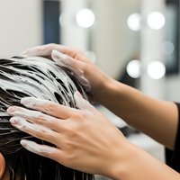 Woman applying white hair mask treatment with hands massaging scalp in salon with bokeh lights background for haircare routine.