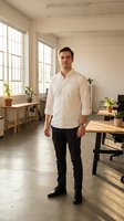 Young Caucasian male professional in casual business attire standing in modern office space with natural light, wooden desks, potted plants, and minimalist decor for corporate workplace concepts.
