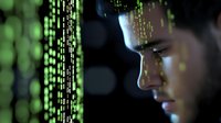 Young Asian male programmer looking at digital code projection in dark room with green matrix style numbers and data streaming on face.
