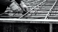 Worker in work gloves tying steel reinforcement bars on concrete foundation framework, construction site detail in black and white.