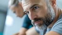 Mature bearded Caucasian man with gray hair and confident expression wearing casual t-shirt, close-up portrait with blurred background and soft lighting.