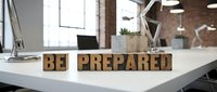 Wooden block letters spelling "BE PREPARED" on white desk in modern office workspace with brick walls and natural lighting.
