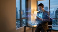 Young Asian businessman analyzing financial data on smartphone with holographic charts in modern office overlooking city skyline at dusk.