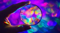 Holographic glass lens held against vibrant neon bokeh background showing rainbow spectrum colors and light refraction patterns in macro photography.