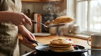 Person in cozy sweater flipping golden pancakes in warm kitchen with steam rising from hot breakfast stack on cast iron pan during morning cooking.