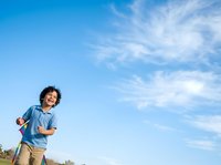 Young boy with curly dark hair and medium skin tone running outdoors against blue sky with white clouds. Child wearing casual denim shirt and khaki pants, enjoying active play in open field during daytime.