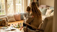 Mother holding toddler son in cozy living room with toys scattered on floor, warm natural lighting through windows creating intimate family moment at home.
