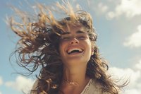Young Caucasian woman laughing with windswept hair against blue sky with white clouds, expressing pure joy and freedom in natural sunlight, lifestyle portrait.