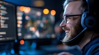Caucasian male programmer with headset smiling while working on code at night, illuminated by multiple computer screens in dark office environment.
