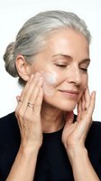 Mature Caucasian woman applying facial cream with gentle massage motions for anti-aging skincare routine on white background.
