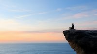 Silhouette of person sitting alone on cliff edge overlooking calm ocean at sunset, peaceful meditation moment against pastel colored sky and horizon.