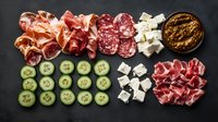 Appetizing arrangement of cured meats, fresh cucumber slices, feta cheese cubes and pesto sauce on dark background, overhead view of Mediterranean antipasti platter.