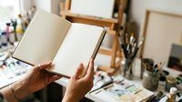 Open notebook with blank pages held by hands in artist studio workspace with easel, paintbrushes, and supplies, suitable for creative projects and art education.