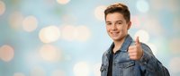 Happy young Caucasian male teenager in denim jacket giving thumbs up gesture against soft bokeh background for positive lifestyle and success concepts.