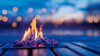 Glowing campfire with bright flames burning on wooden deck at twilight with bokeh lights reflecting on water in background creating cozy outdoor atmosphere.