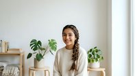 Young woman with long braided hair and medium skin tone smiling at camera in bright room surrounded by potted green plants and wooden shelving, ideal for lifestyle, wellness, and home decor content.