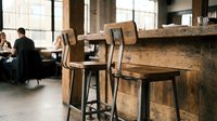 Wooden bar stools at rustic counter in modern loft cafe with industrial windows and blurred customers in background, suitable for hospitality and interior design projects.