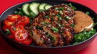 Grilled chicken breast with sesame crust served on fresh greens with sliced tomatoes, cucumber, and creamy sauce in dark plate on red background.