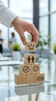 Male hand placing wooden block with crown symbol on stacked cubes showing leadership, teamwork, strategy and management icons in modern office environment with blurred colleagues and windows.