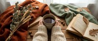 Cozy autumn flat lay with knitted mittens, hot tea, open book and dried eucalyptus branches on soft blankets creating warm seasonal hygge atmosphere.