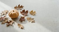 Small orange pumpkin surrounded by scattered brown autumn leaves on neutral linen fabric background for seasonal decoration and harvest themes.