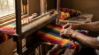 Elderly hands weaving colorful rainbow striped textile on traditional wooden loom in rustic workshop with natural sunlight streaming through window.