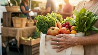 Fresh organic vegetables in reusable paper bag held by farmer at market, including broccoli, carrots, tomatoes and leafy greens for sustainable shopping and healthy eating.