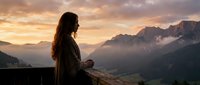 Woman with long dark hair, wearing casual clothing, standing on a wooden railing overlooking misty mountain valley at sunrise or sunset. Contemplative traveler enjoying scenic alpine landscape with golden hour lighting.
