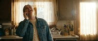 Young Hispanic woman laughing joyfully while talking on smartphone in cozy kitchen with warm sunlight streaming through curtains creating happy domestic moment.